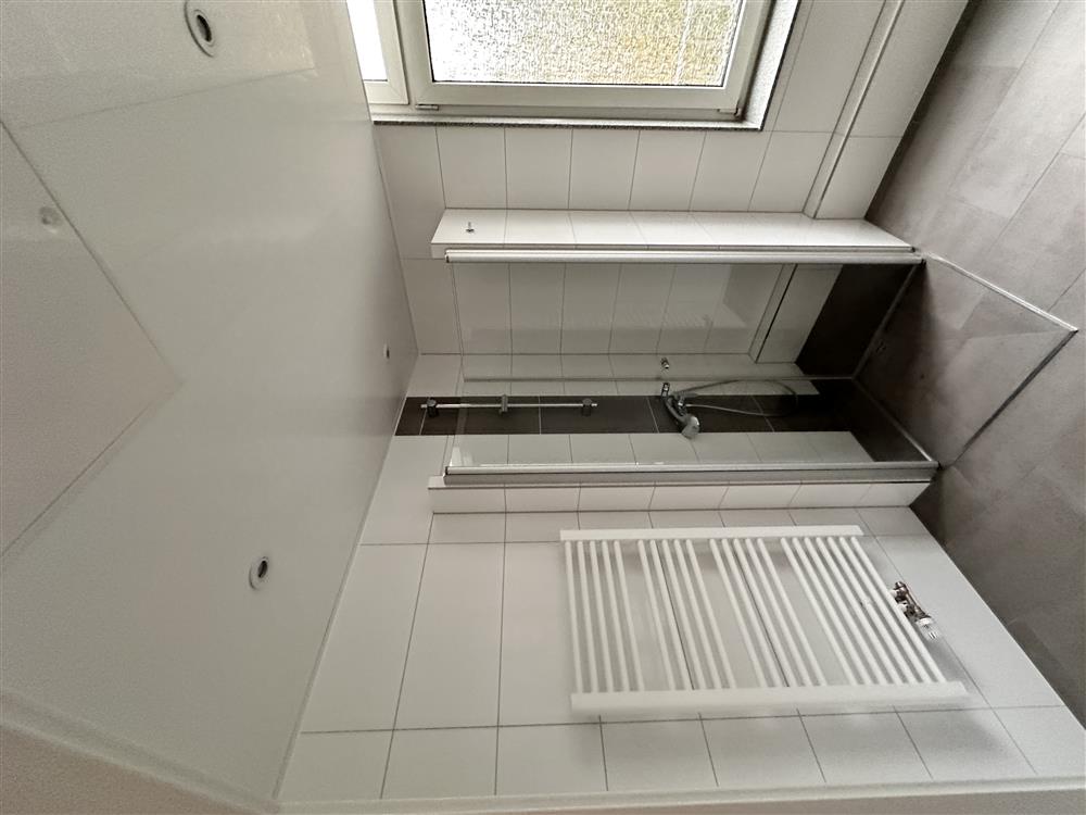 Dusche mit Glasabtrennung in einer modernen, frisch renovierten Wohnungshalle, weiße Wandfliesen und ein Handtuchheizkörper, große Fenster für natürliches Licht, ideale Ausstattung für Komfort und Funktionalität.